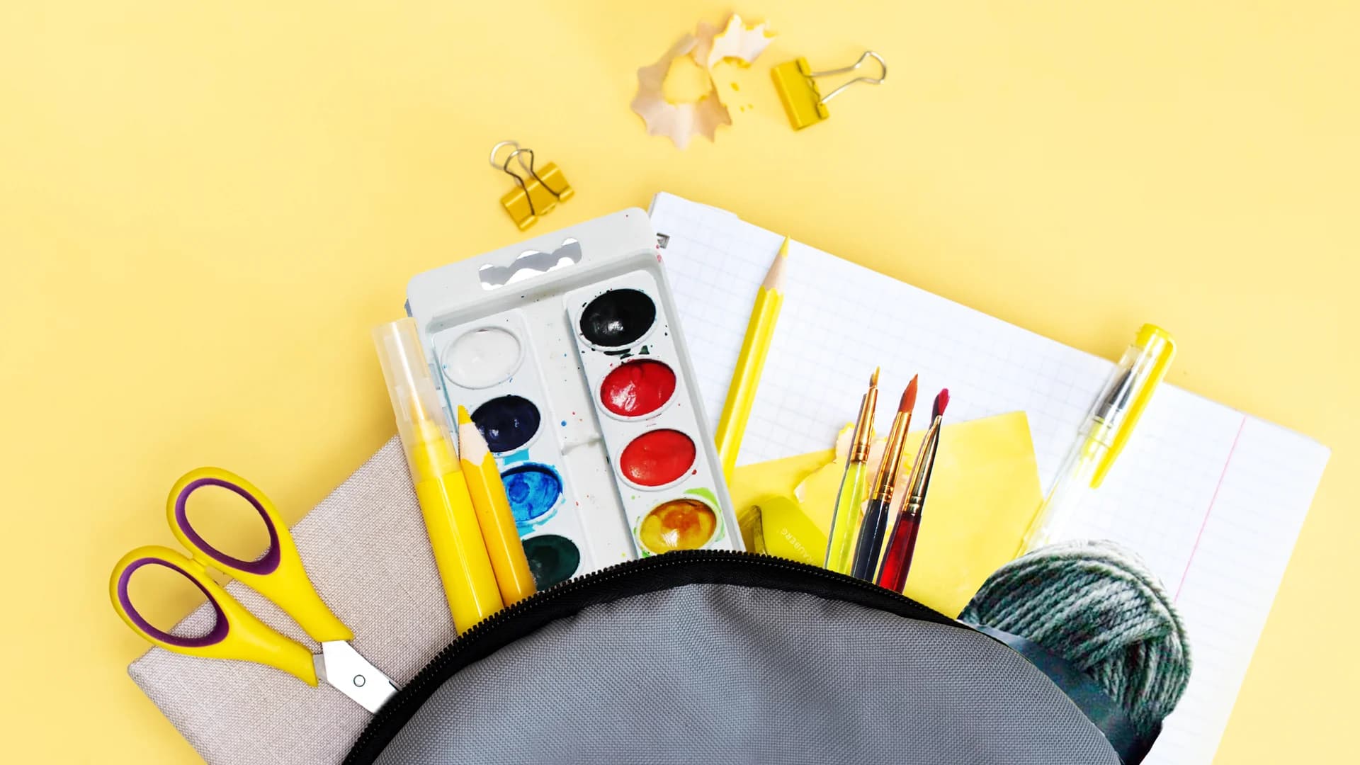 Analog bag contents laid out on yellow background: watercolor paint set, brushes, scissors, notebook, yarn, pencils, and other screen-free creative supplies