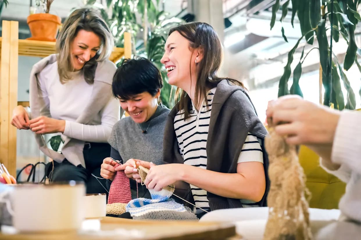 Group of friends laughing and knitting together at a craft workshop, representing analog hobbies and screen-free social activities